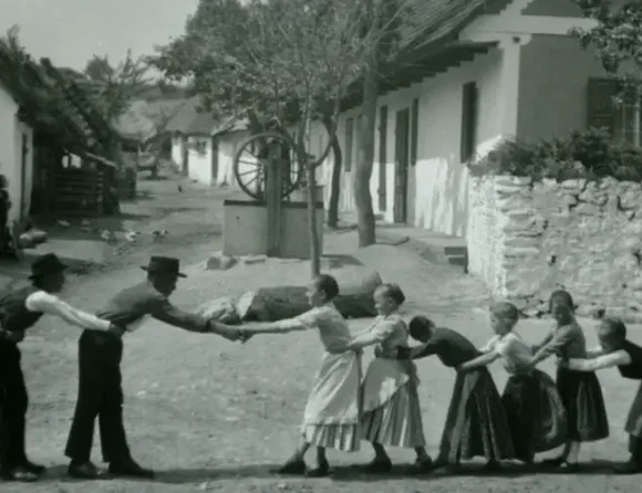 Links stehen die Jungen (evt. auch Männer) in einer Reihe hintereinander und halten jeweils ihren Vordermann an der Hüfte fest; gegenüber eine Reihe Mädchen hinterander, die sich ebenfalls an der Hüfte festhalten. Der vorderste Junge und das vorderste Mädchen stehen sich gegenüber und halten sich an den Händen; im Hintergrund steht ein Mädchen, ihre Arme um einen Baum geschlungen; einige Häuser des Dorfes, Mauern, ein Zaun und eine Straße sichtbar