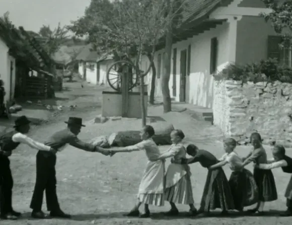 Links stehen die Jungen (evt. auch Männer) in einer Reihe hintereinander und halten jeweils ihren Vordermann an der Hüfte fest; gegenüber eine Reihe Mädchen hinterander, die sich ebenfalls an der Hüfte festhalten. Der vorderste Junge und das vorderste Mädchen stehen sich gegenüber und halten sich an den Händen; im Hintergrund steht ein Mädchen, ihre Arme um einen Baum geschlungen; einige Häuser des Dorfes, Mauern, ein Zaun und eine Straße sichtbar