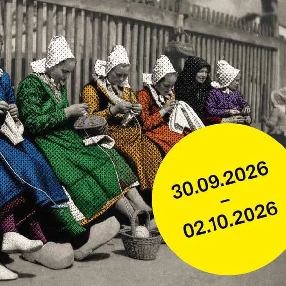 In einer Reihe im Freien sitzende Frauen in Trachten stricken. Die Schwarz-Weiß-Fotografie ist nachkoloriert.