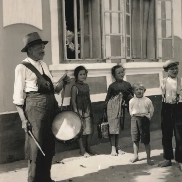 Ein älterer Mann mit einer Trommel vor einem Haus. Um ihn herum stehen Kinder.
