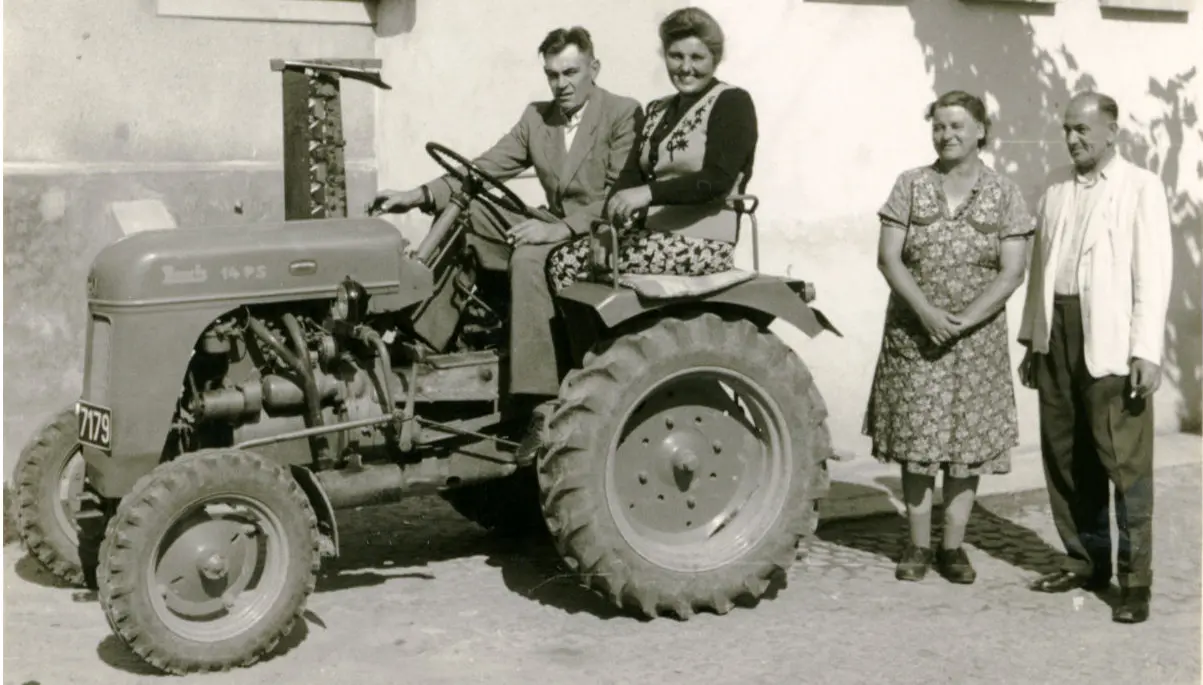 Ein Mann und eine Frau sitzen auf einem Traktor vor einem Haus, daneben stehen ein weiterer Mann und eine Frau.