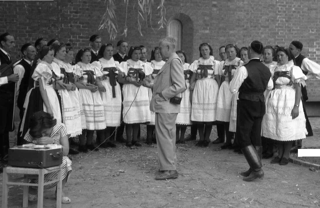 Ein Mann mit einem Mikrofon nimmt einen Chor von Frauen in Tracht auf.
